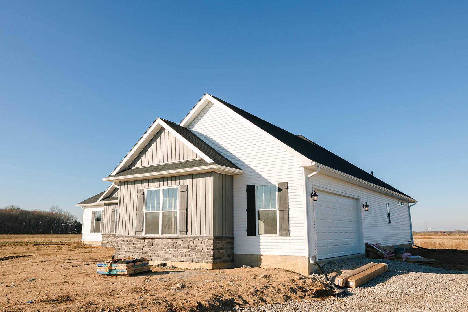 Framed house under construction with exposed plywood siding, attached garage, white-framed window, stack of cement blocks on grassy lot, under clear blue sky