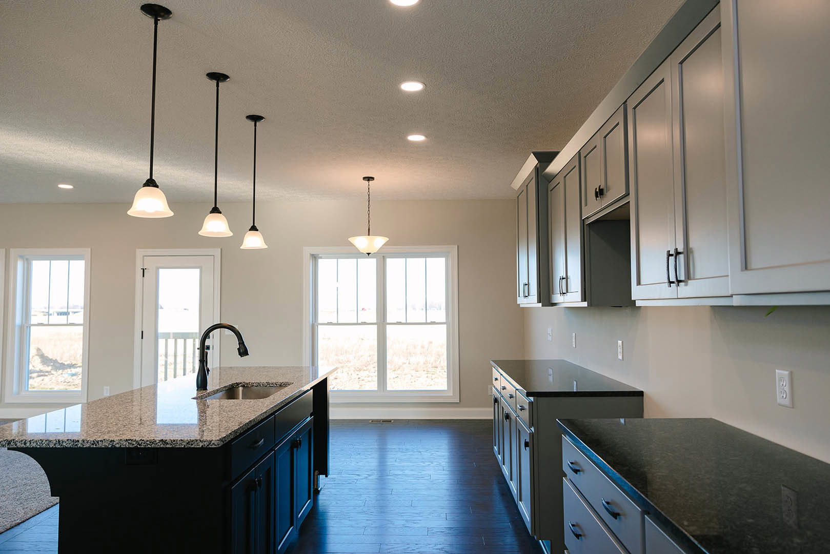 Spacious kitchen featuring a large central island with granite countertops, built-in sink, white cabinetry, stainless steel appliances, pendant lighting, and a window providing