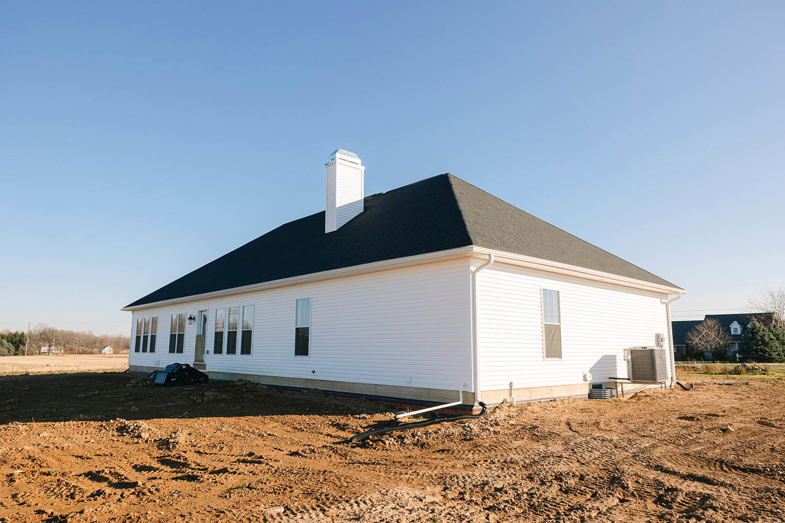 White siding house with black shingle roof, prominent chimney, multiple windows, and landscaped front yard under clear sky