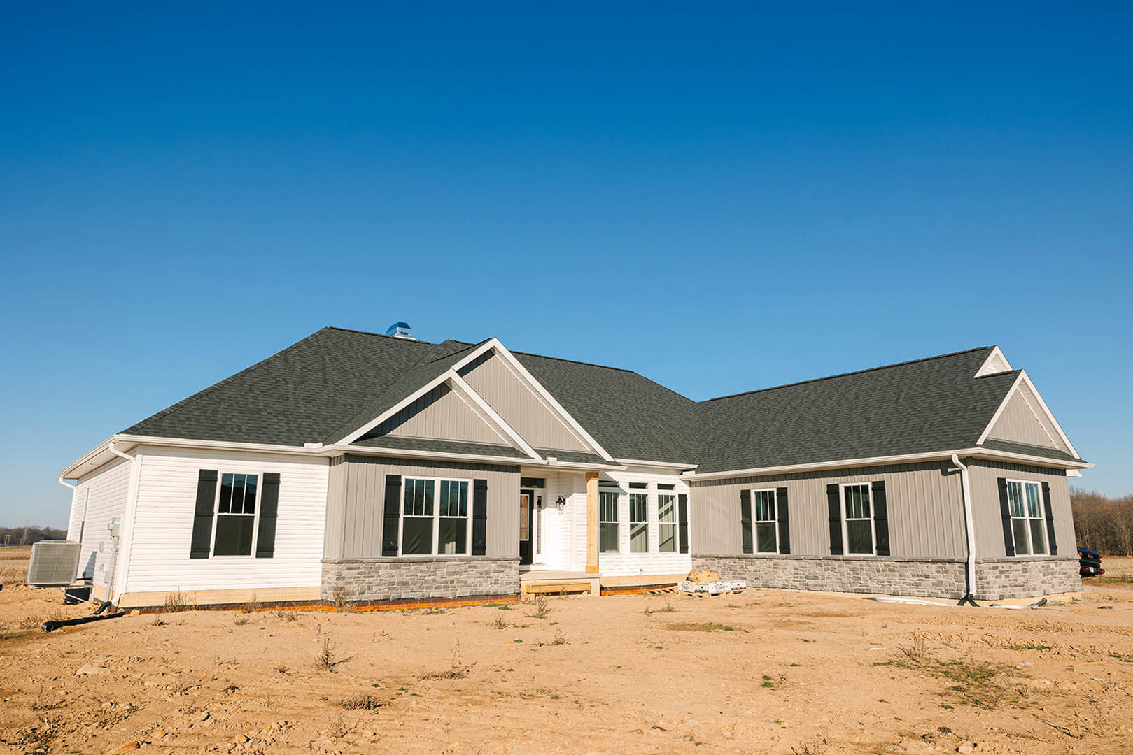 Framed house under construction with exposed wooden beams, dirt lot in foreground, and clear blue sky overhead