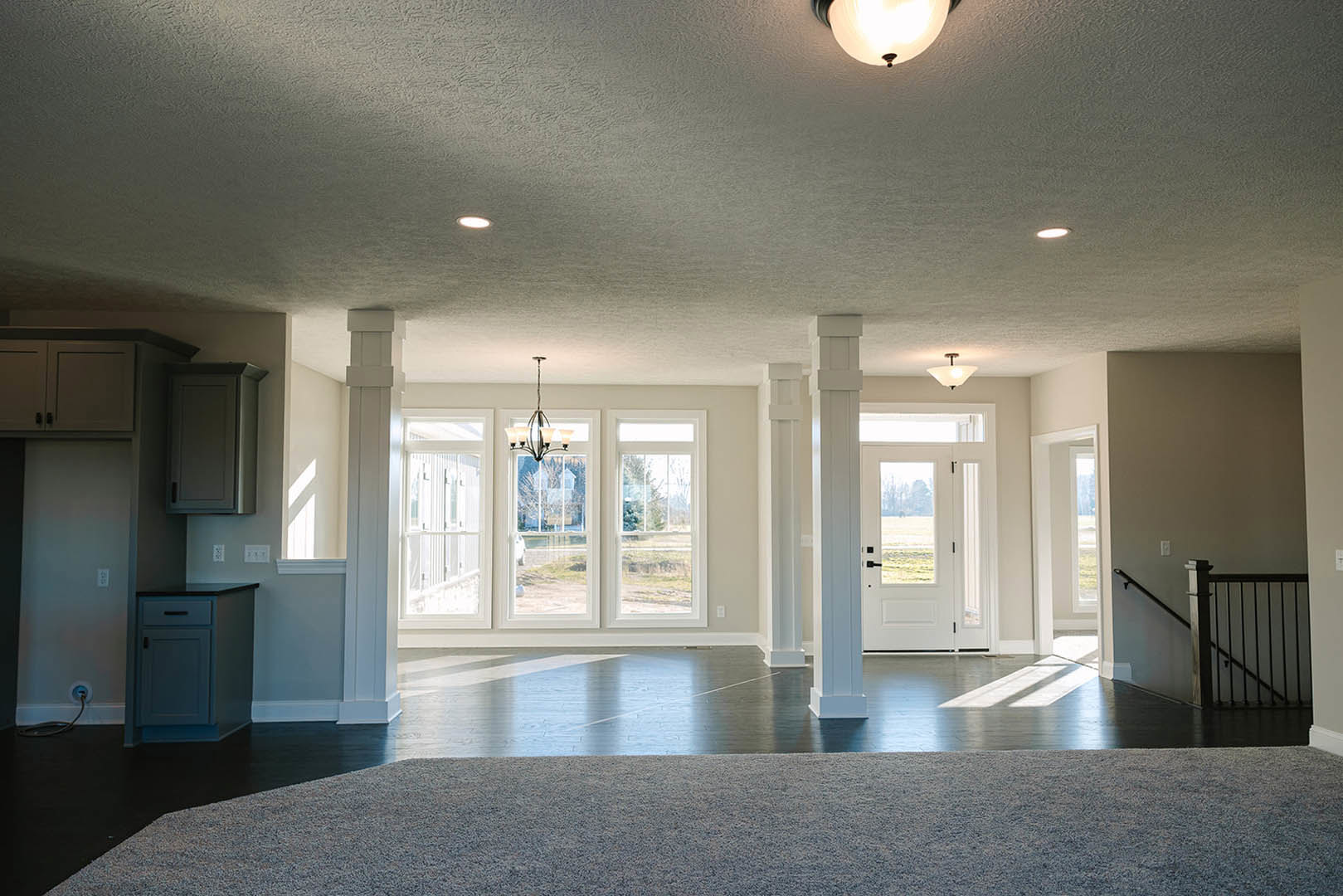 Spacious room featuring a large window, white door, plush carpet flooring, modern light fixture with white shade, staircase with sleek metal railing, and built-in cabinetry with