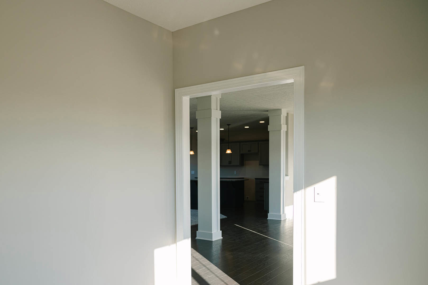 Wide doorway framed by white pillars, dark wood flooring with white trim, soft overhead lighting, plaster walls, and ceiling in a modern residential interior.