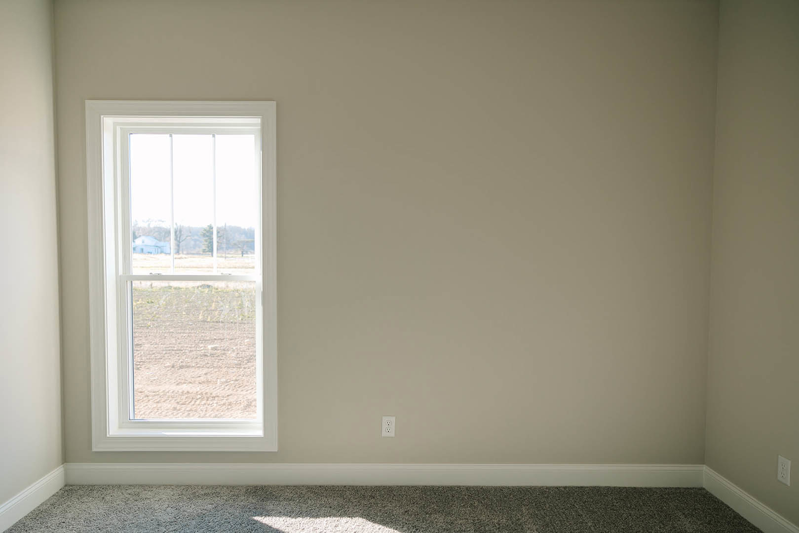 Person standing on light carpeted floor in a bathroom with tile walls, large window overlooking a grassy field and neighboring house, window blinds partially drawn