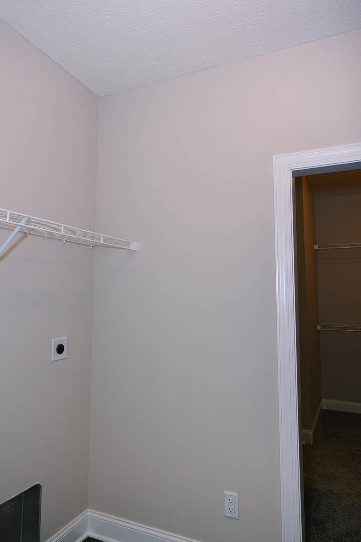 White closet with built-in shelf, white plaster walls, black carpet flooring, and a black round wall fixture near a door.