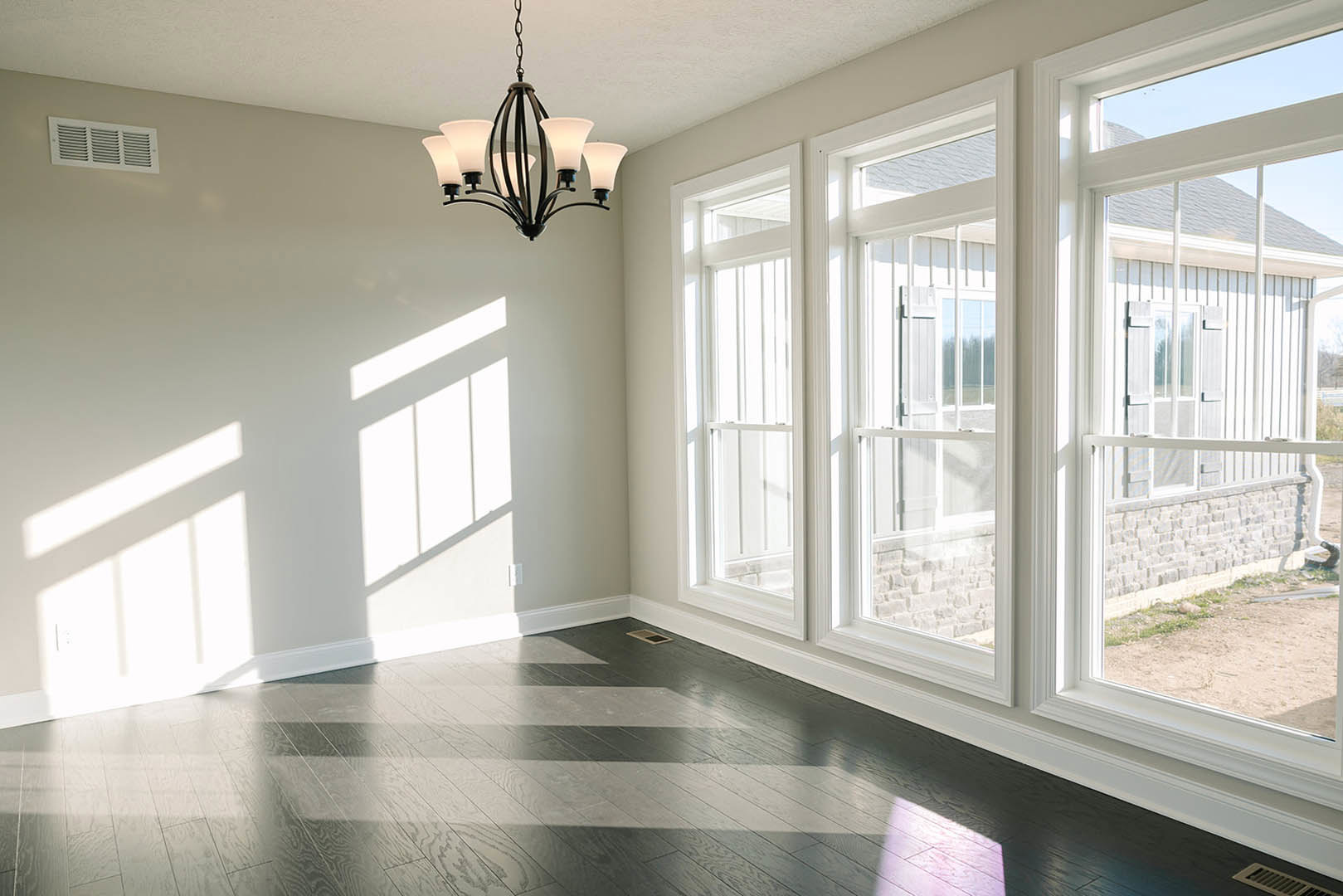 White-walled room with large windows, black and white striped flooring, chandelier, wall vent, and lamp; view of neighboring house through windows