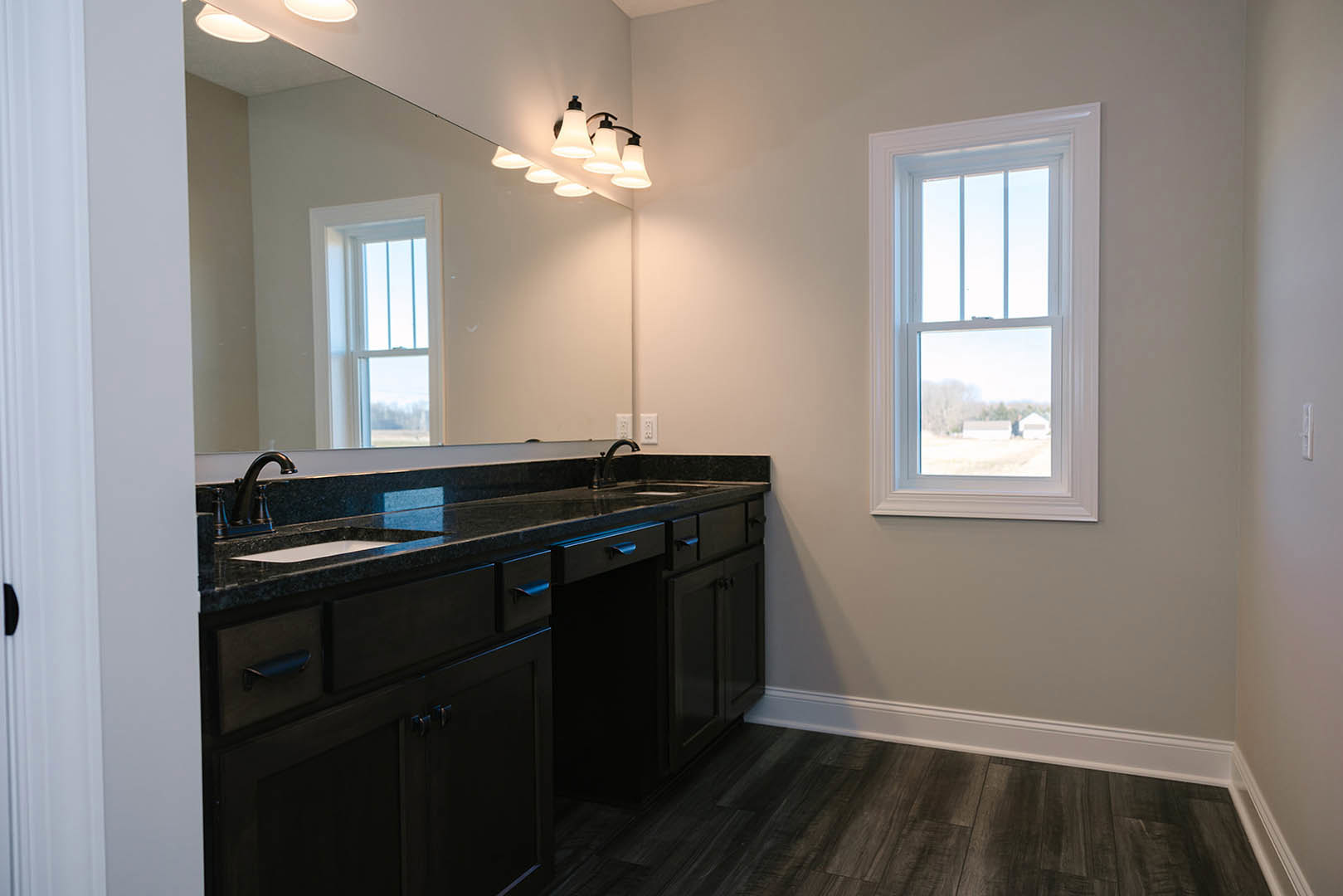 Bathroom with expansive mirror above black countertops, wood flooring with white trim, modern ceiling lights, chrome faucet, and window offering view of neighboring house.