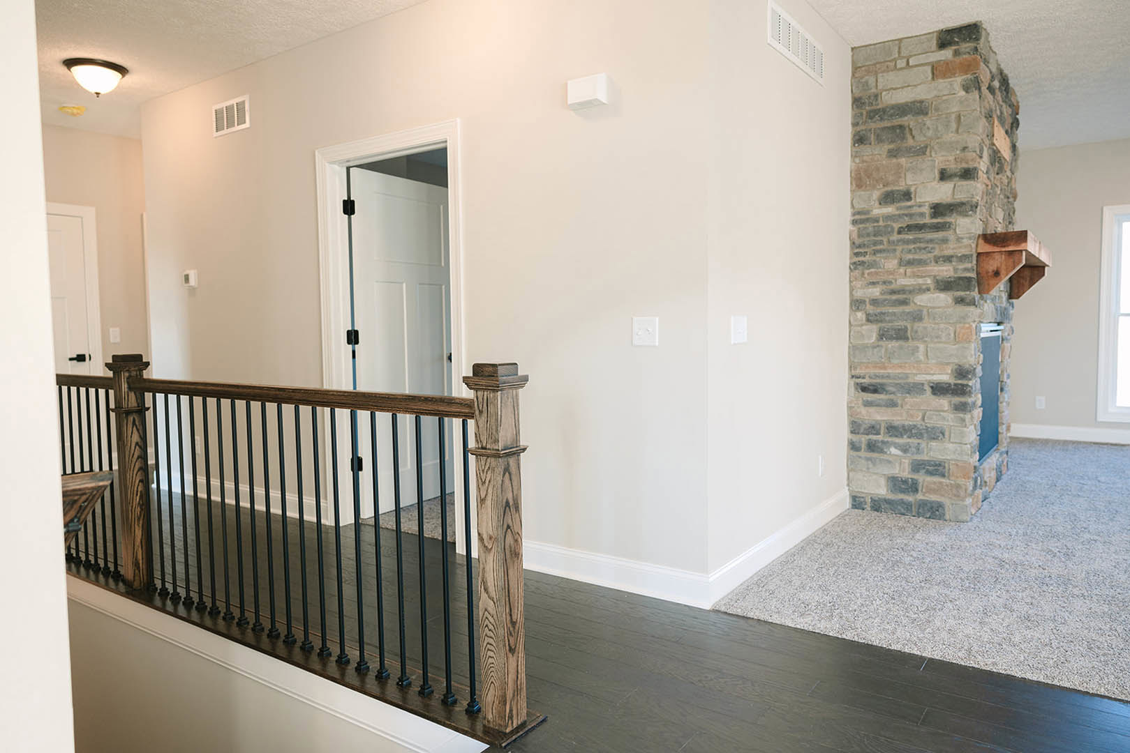 Wood staircase with black metal railing beside exposed brick fireplace, white vent and electrical box on plaster wall, hardwood flooring throughout.