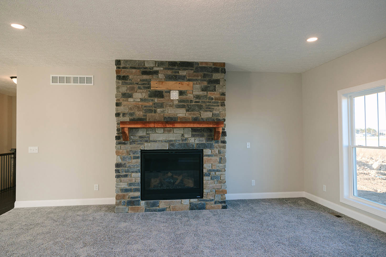 Black-framed fireplace set in a brick accent wall with a wood mantel, large window overlooking a grassy field, plush carpeted floor in a cozy den.