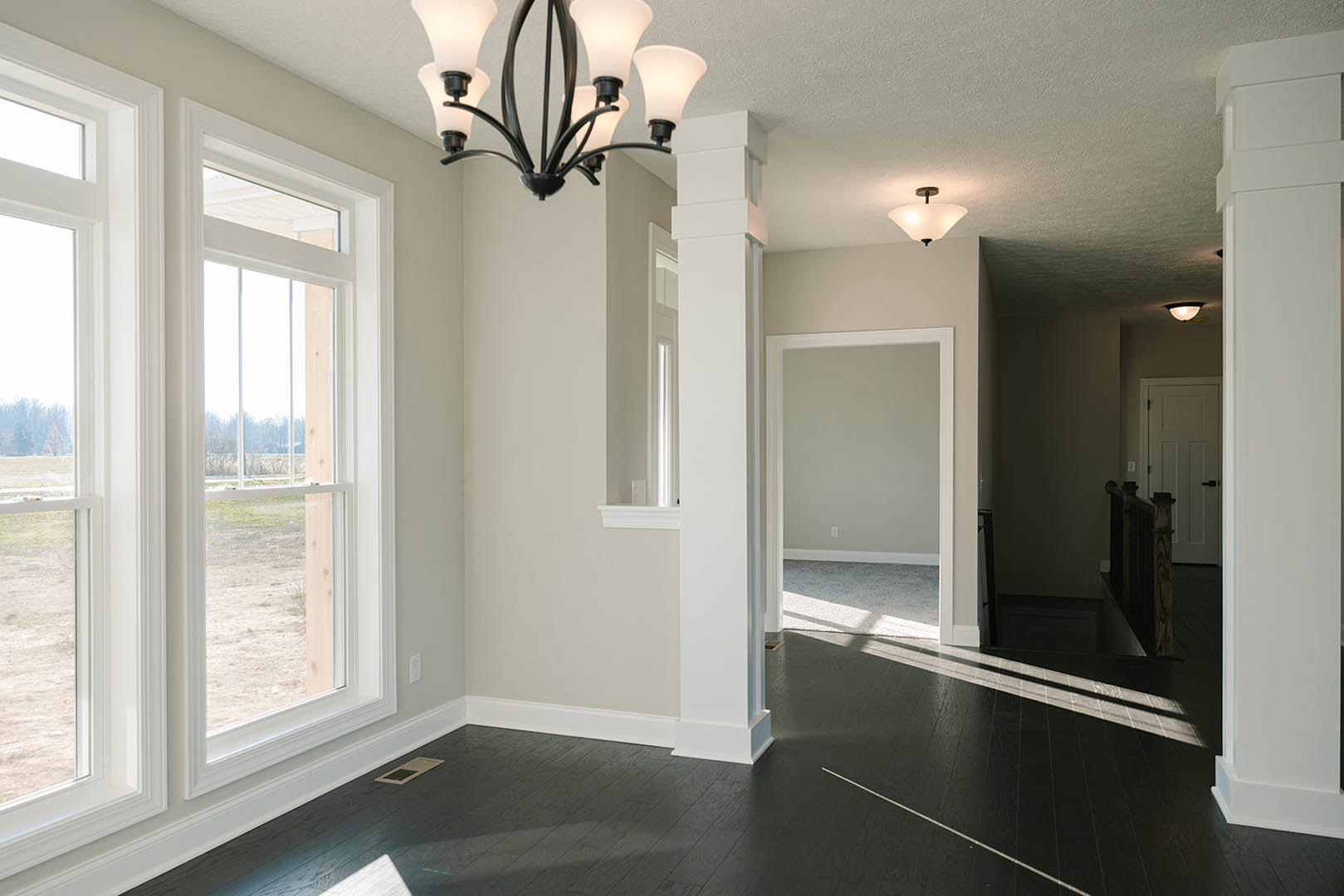Black hardwood floor, white plaster walls, modern chandelier, white door with black handles, large window overlooking grassy field, simple ceiling molding