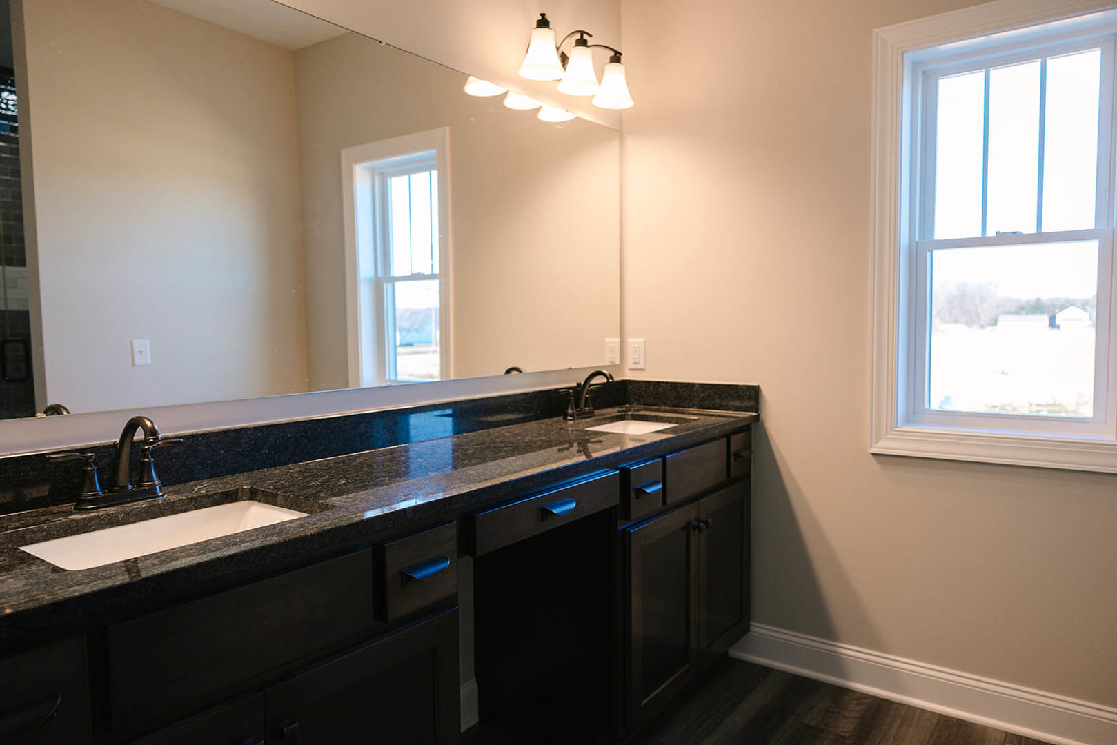 Bathroom with expansive mirror above a wide vanity, modern faucet and sink, light fixture mounted on wall, window overlooking open field, white cabinetry and neutral countertops