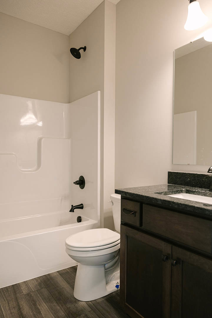 Modern bathroom featuring white ceramic sink and toilet, light gray tile flooring, wood vanity cabinet, wall-mounted black faucet, rectangular mirror, and contemporary sconce lamp.