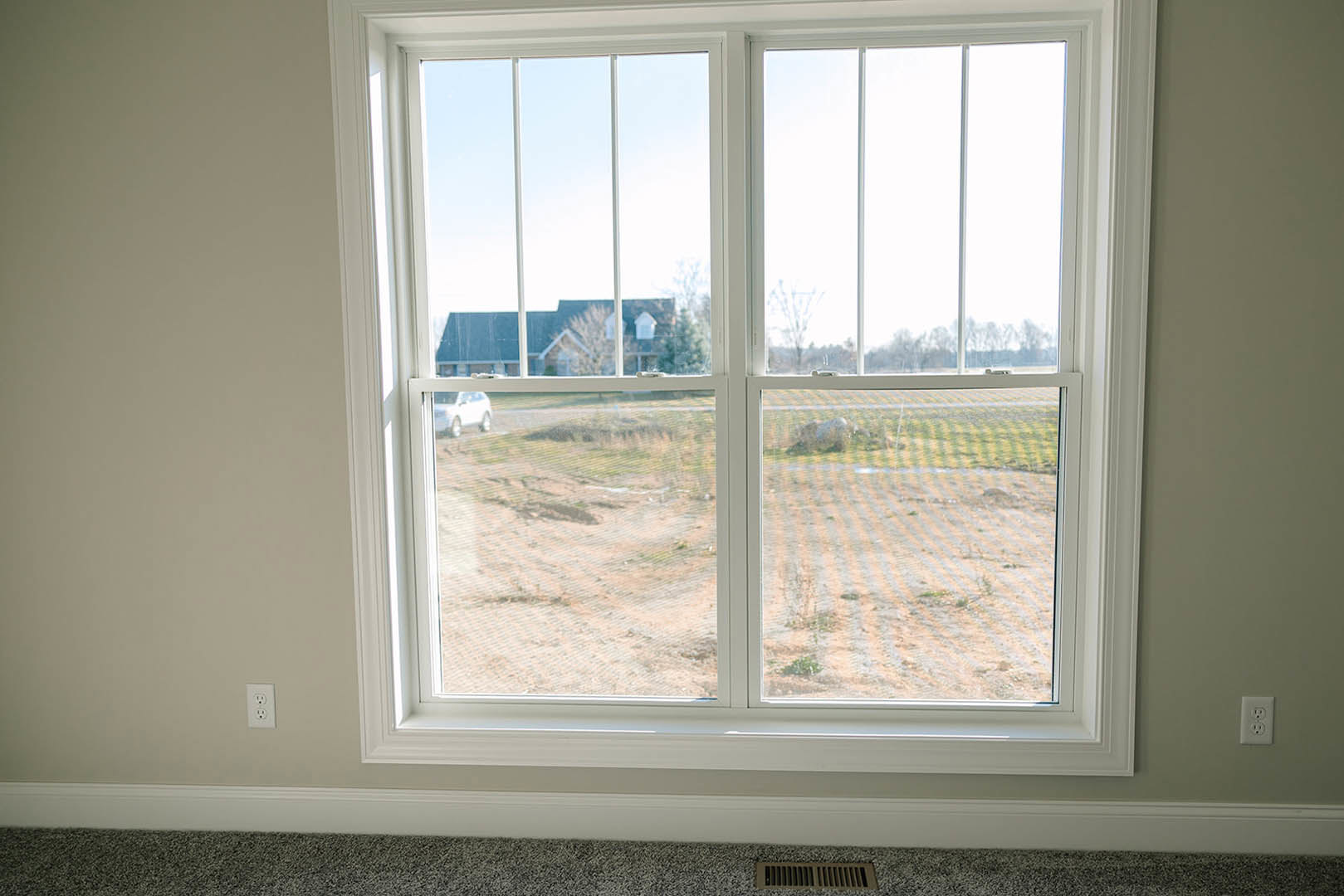 Large window with white trim in a sunlit room, offering a view of a grassy field and distant house; nearby electrical outlet visible on pale wall, window blinds partially drawn.