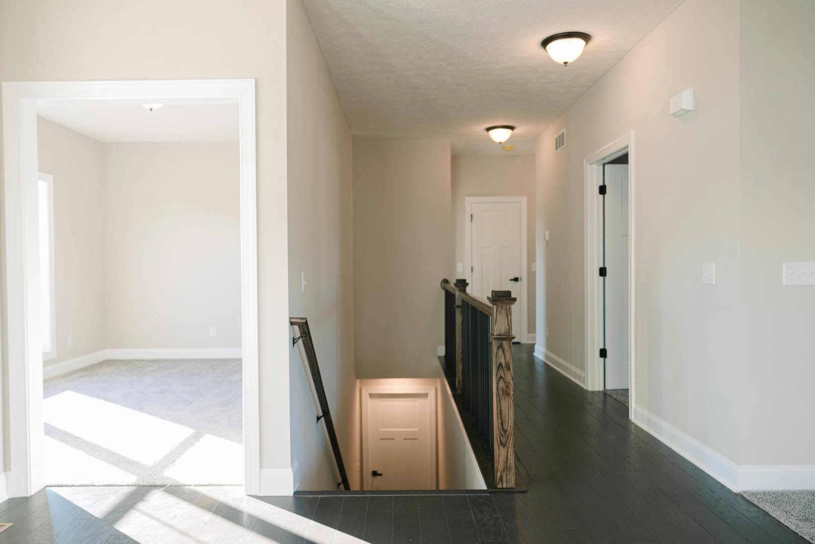 Hallway with light wood flooring, white plaster walls, open white door with black handle, wooden staircase with vertical posts, round ceiling light fixture