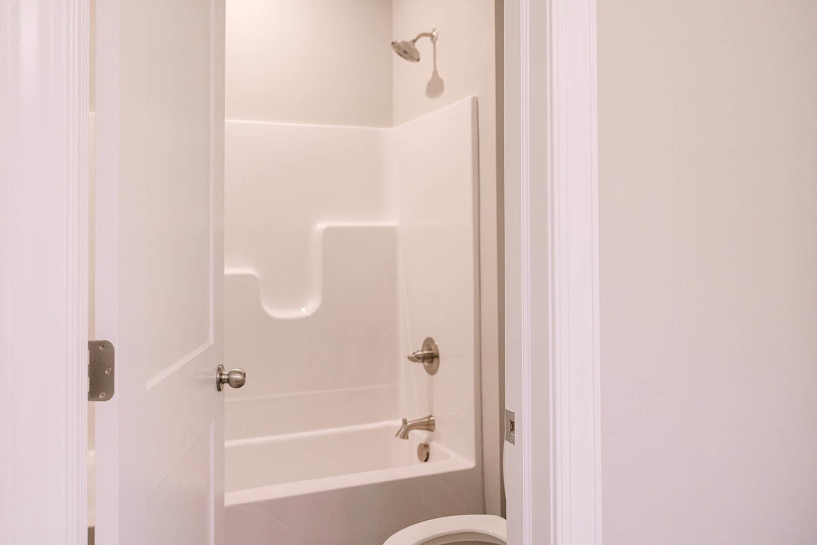 White bathtub with silver faucets beside a white toilet, light tile flooring, partial view of door with silver handle, blurred faucet and door knob in foreground