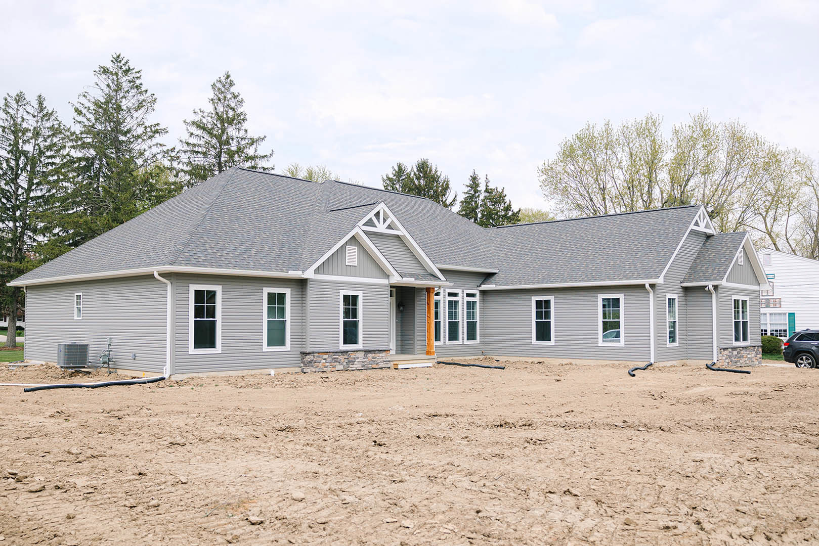 Partially built house with grey roof and white-framed windows, surrounded by dirt field, construction materials, and a large white box with metal bars in foreground
