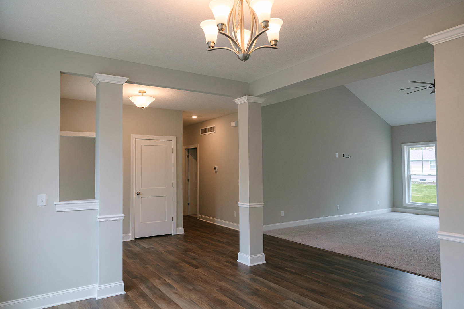 Open living space featuring warm wood flooring, ornate chandelier, white walls with crown molding, black-handled door, and large window overlooking a neighboring white house.