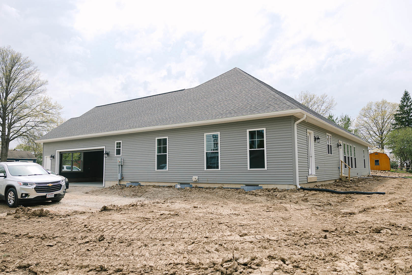 Two-story house under construction with exposed framing, white window trim, dirt yard, silver car parked in front, leafy tree on the side, cloudy sky overhead