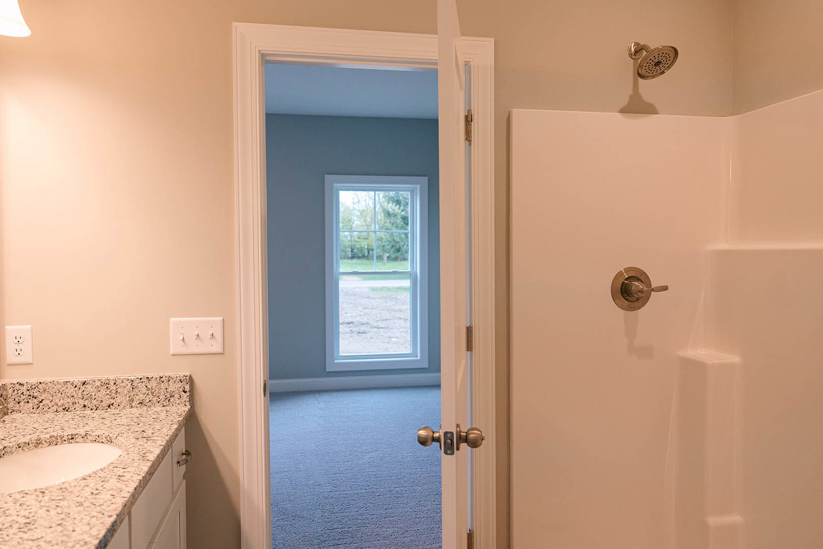 Bathroom with glass shower enclosure, white tile walls, wooden door, marble countertop with round sink, chrome faucet, electrical outlet, and window overlooking grassy field.