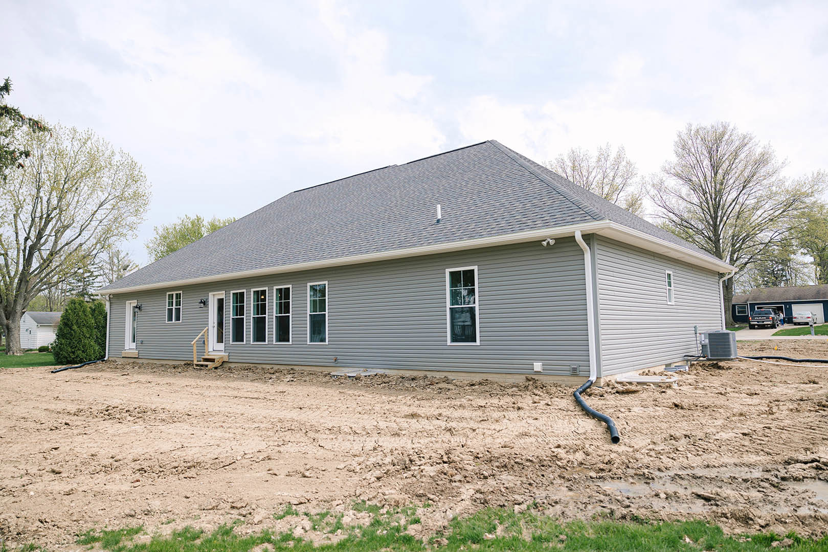 Two-story house under construction with exposed framing, white window frames, dirt yard, surrounding trees, black truck parked in open garage, and air conditioning unit visible