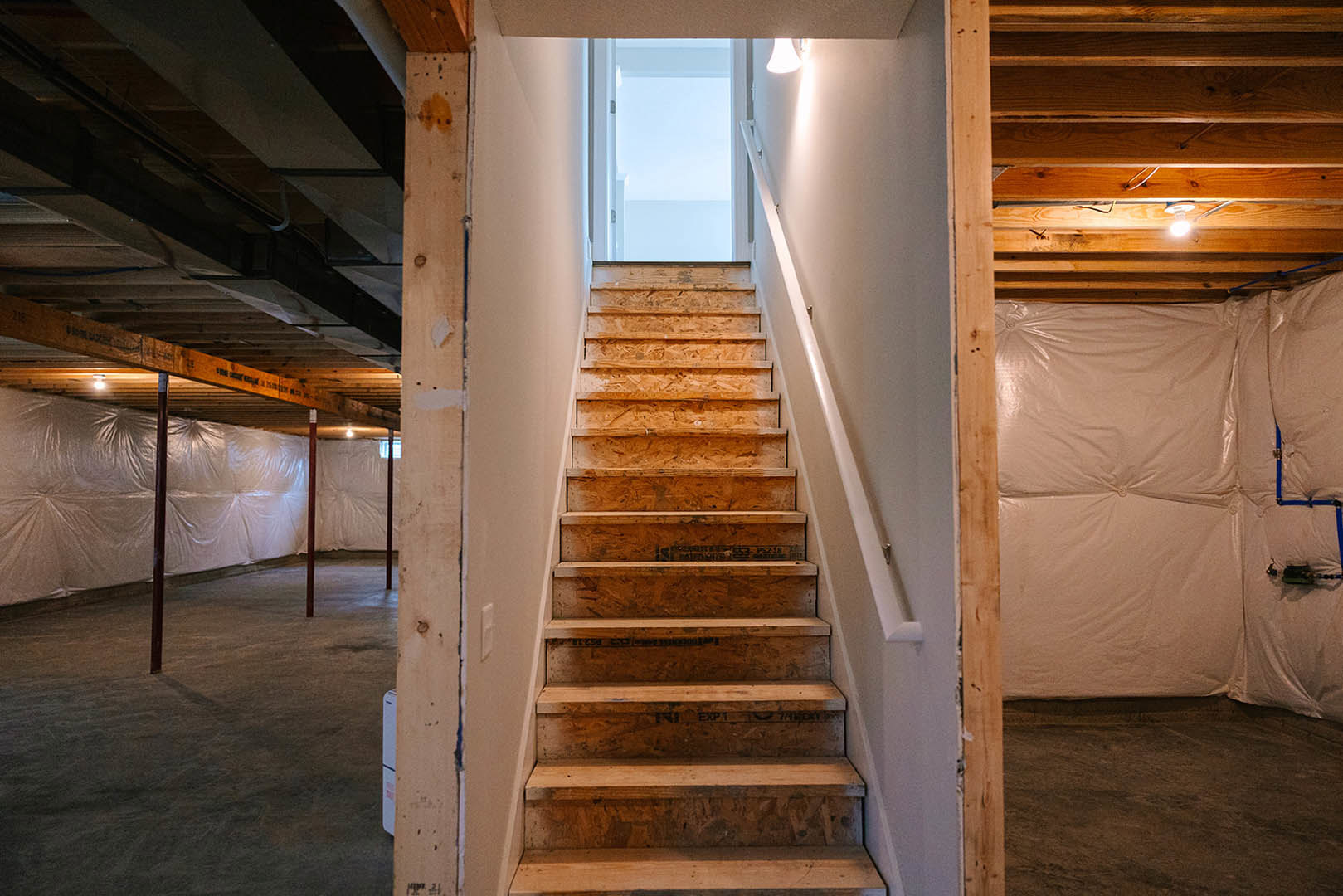Modern staircase with plywood side panels, exposed wood beams, white plastic sheeting covering one wall, close-up of a window and recessed light, unfinished plaster and lumber