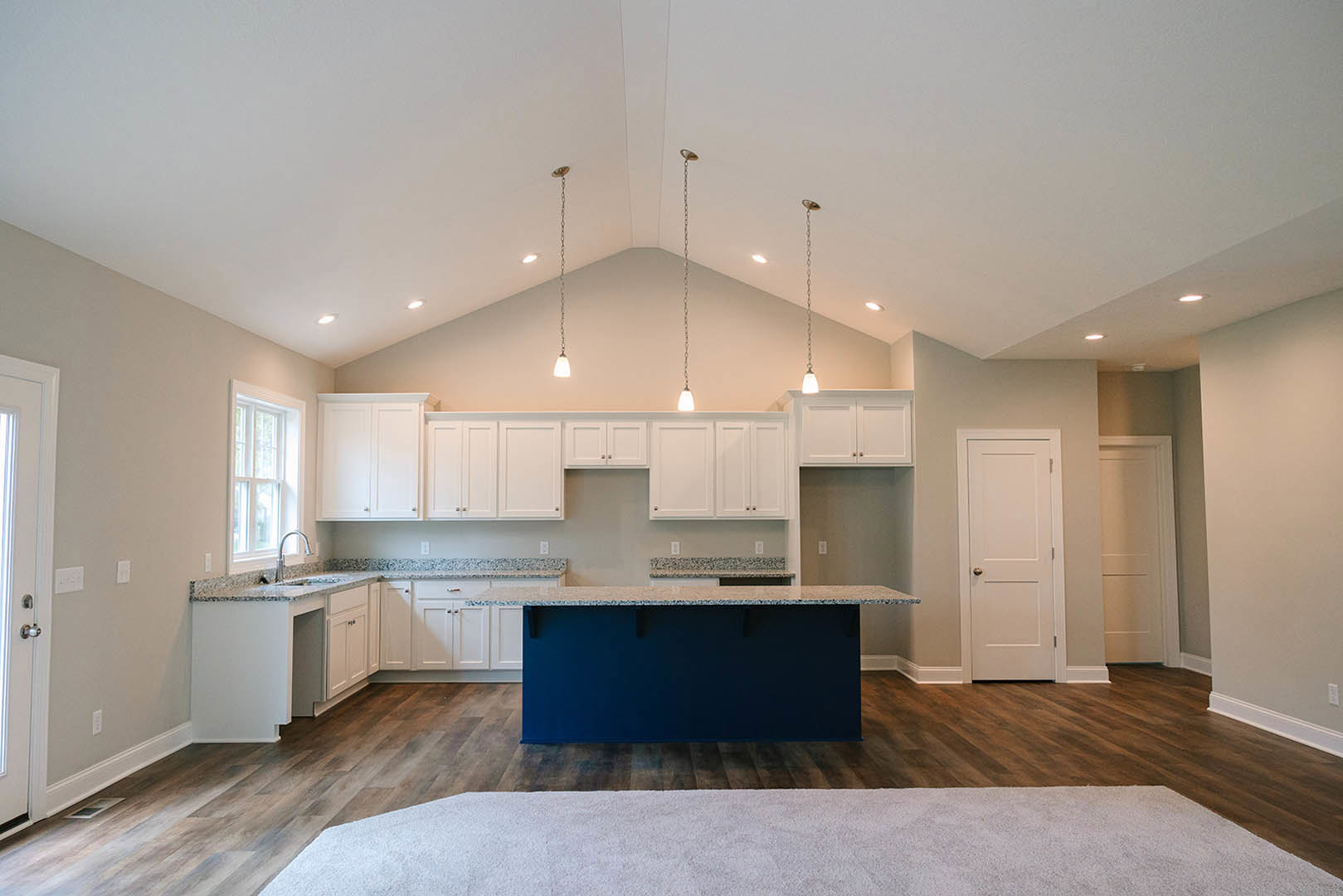Blue kitchen island with white quartz countertop, surrounded by white shaker cabinets, stainless steel sink, and silver hardware; white walls and door, light carpet flooring, large