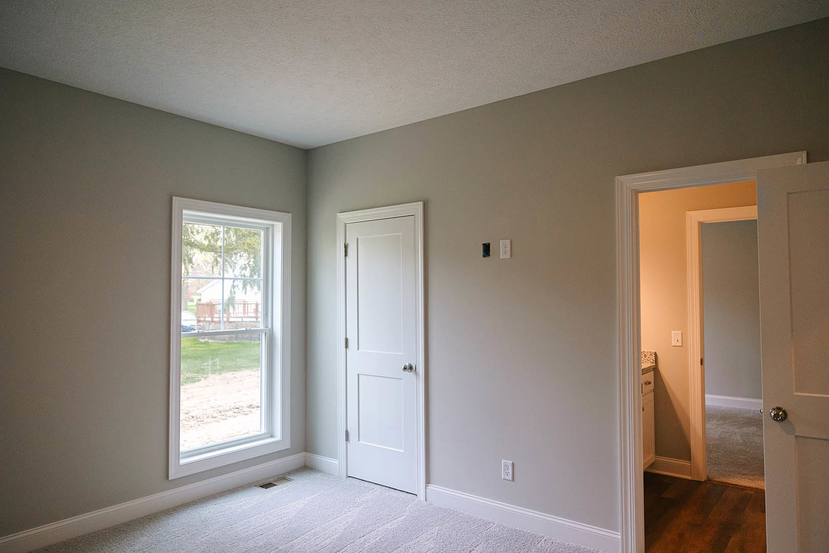 Neutral-toned room with two white doors featuring silver handles, large window overlooking a grassy yard and neighboring house, white carpet flooring with a floor vent