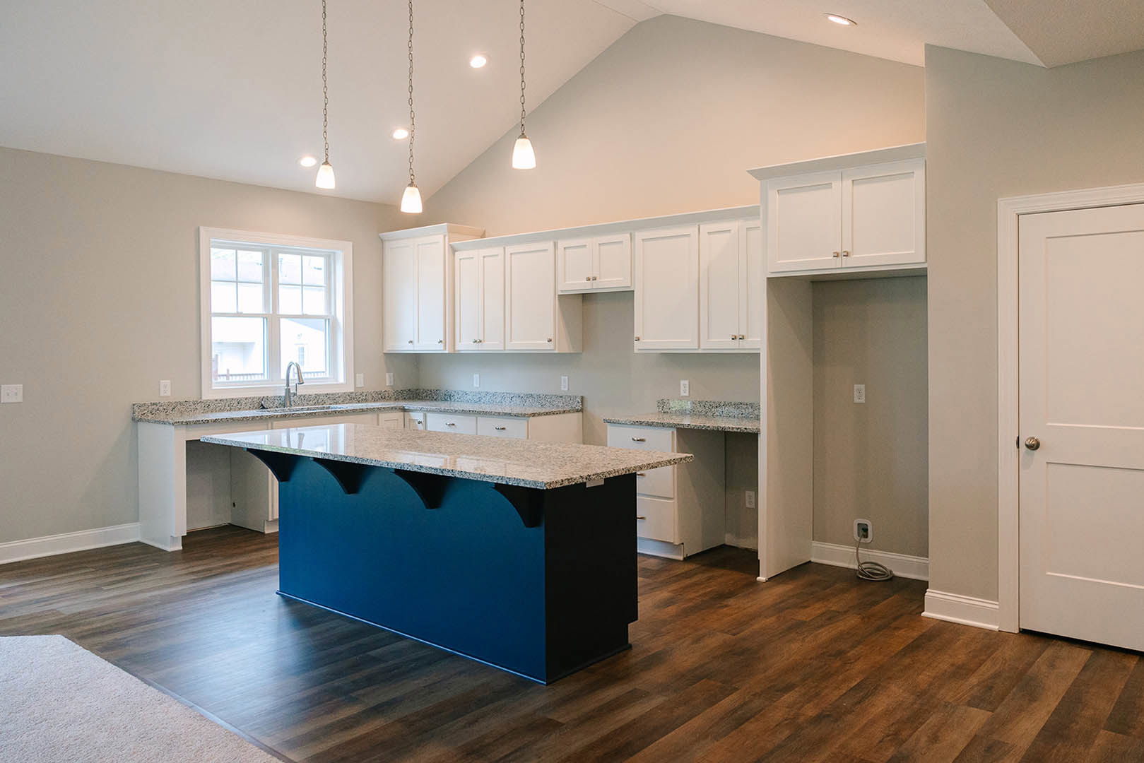 Blue kitchen island with white cabinets, white tile backsplash, stainless steel sink beneath a window, light wood flooring, and white walls with a door and electrical outlet.