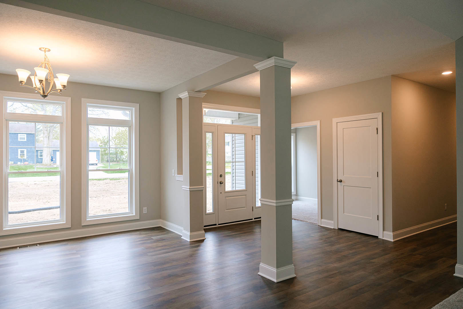 Hardwood floor with white pillars, white doors, and a chandelier; large windows reveal a blue house and tree outside