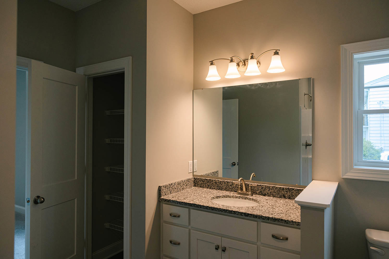 Bathroom with white pedestal sink, rectangular mirror, two-light wall fixture, white framed window, visible closet shelf, and close-up of toilet; neutral tile walls and cabinetry.