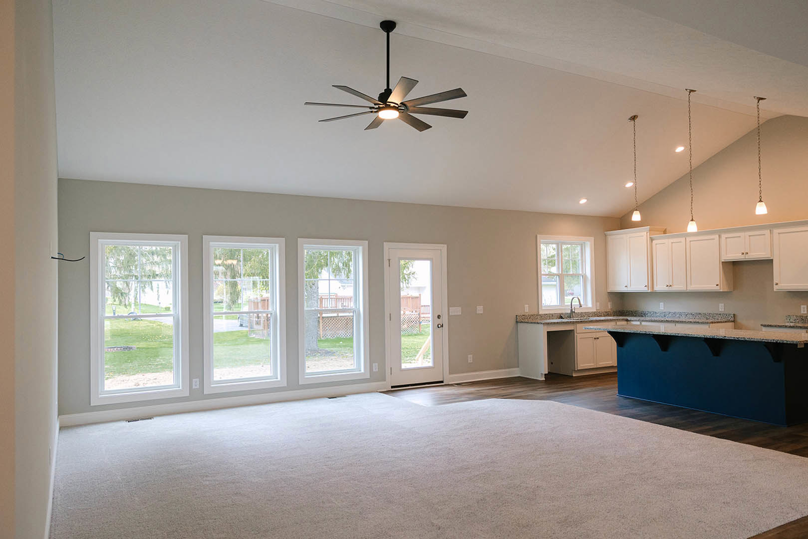 Spacious living room with carpet flooring, open kitchen featuring blue accent wall and countertop, large ceiling fan with light fixture, glass door and window overlooking green