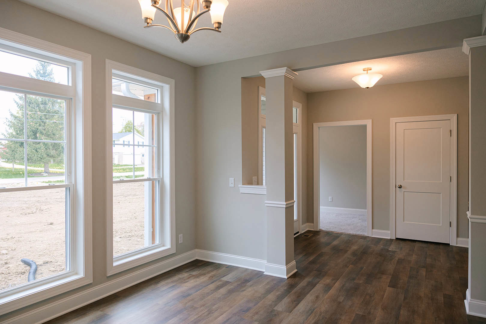 Wood floor room with white walls, decorative molding, silver door knob on white door, ceiling chandelier, and window overlooking neighboring house
