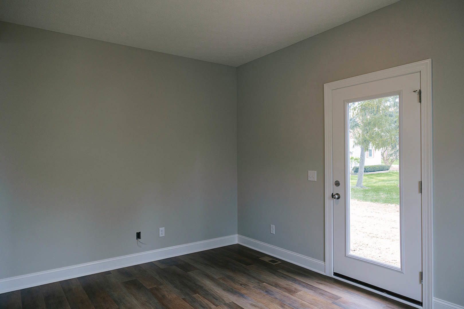 Wood flooring with white baseboards, white walls, a solid door, and a glass door opening to a lawn with a tree visible outside; white ceiling features a black stripe.