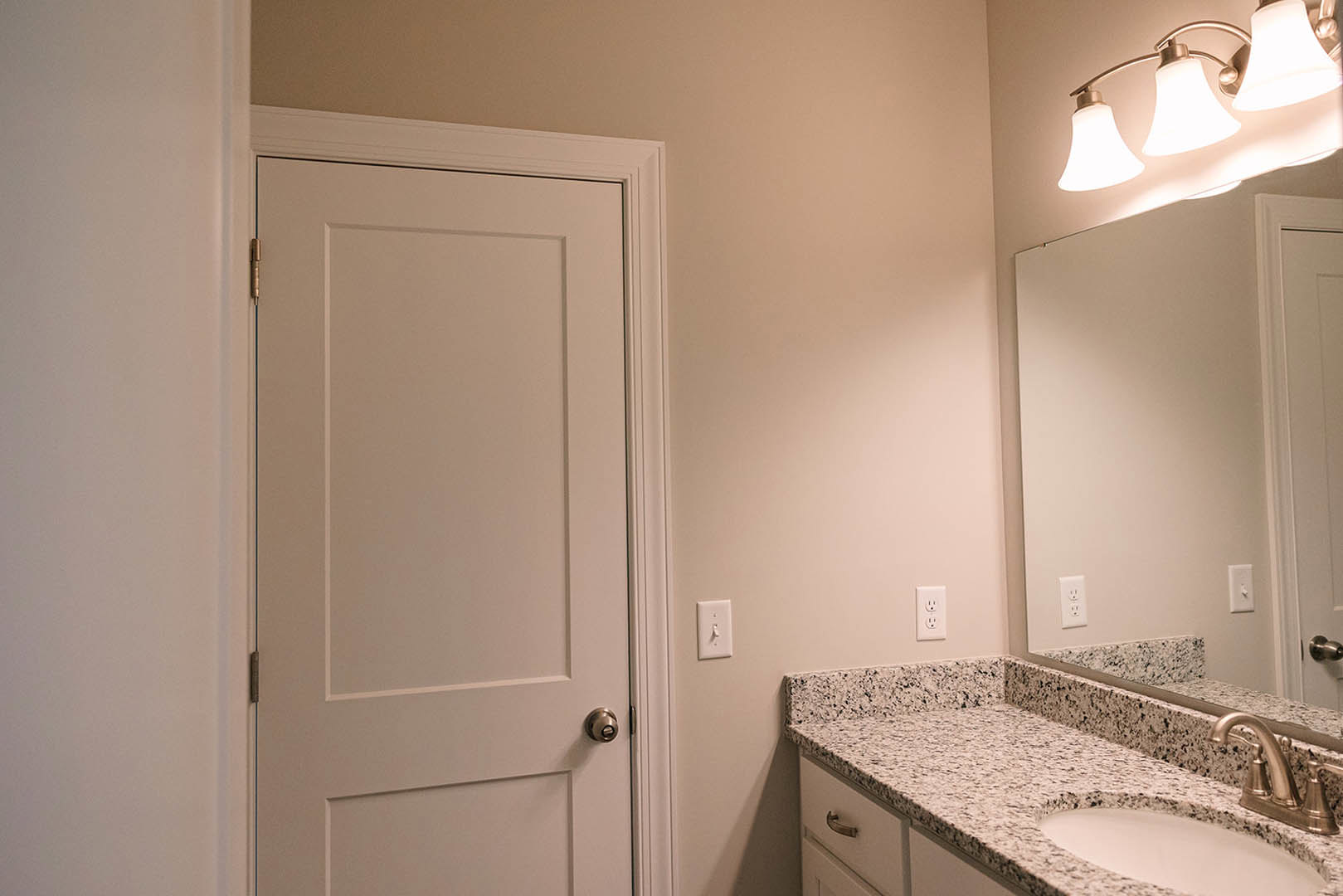 Modern bathroom with white tile walls, rectangular mirror above a sleek sink, chrome faucet, light fixture mounted above the mirror, white countertop, and a white door with metal