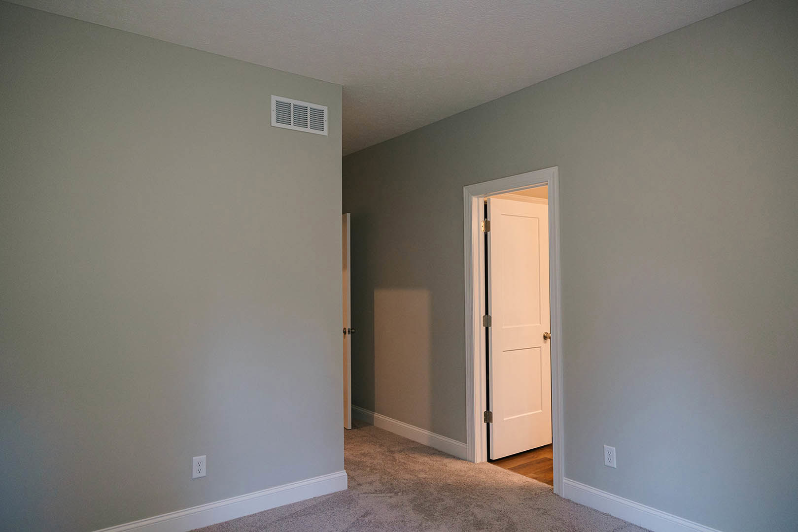 White hallway with plush beige carpet, open white door featuring gold knob, wall-mounted vent, smooth white walls and ceiling