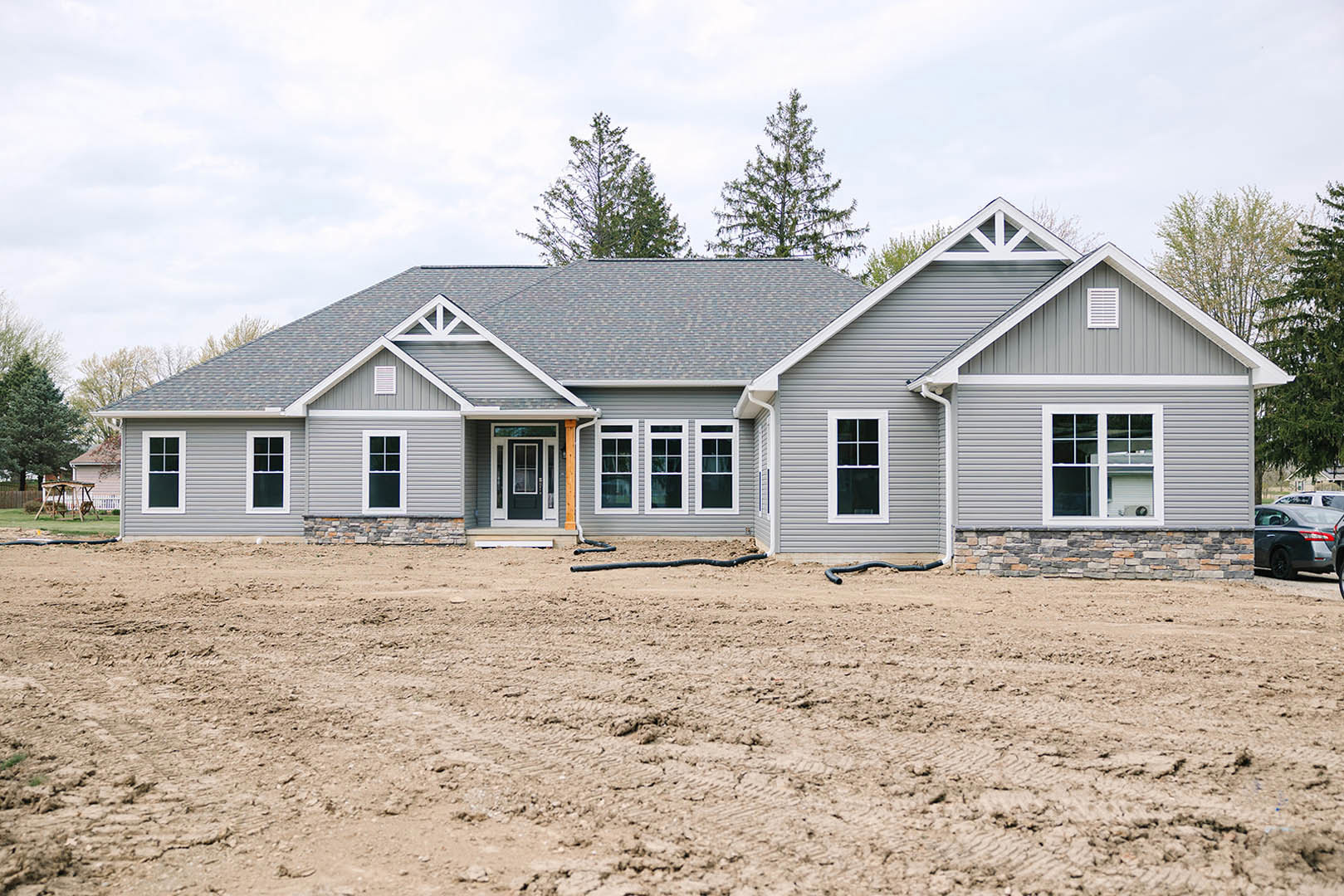 Grey-roofed house under construction with white-framed windows, dirt ground scattered with black hoses, man standing beside a car, cloudy sky and trees in background