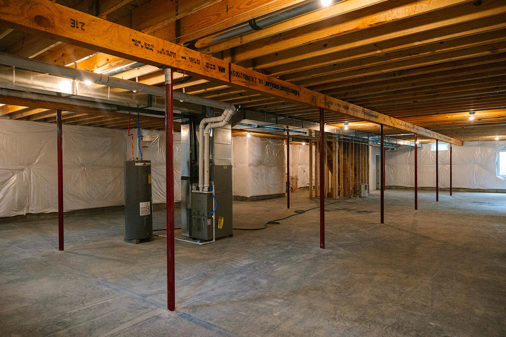 Basement room with exposed wooden ceiling beams, concrete floor, black metal cylinder, visible pipes along wall, and white sheet draped over a pole.