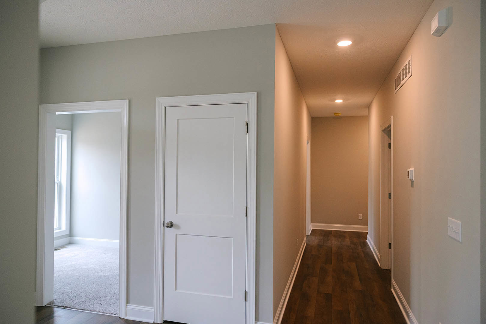 Hallway with dark wood flooring, white door with silver knob, wall vent, light switch, and adjacent room featuring a window