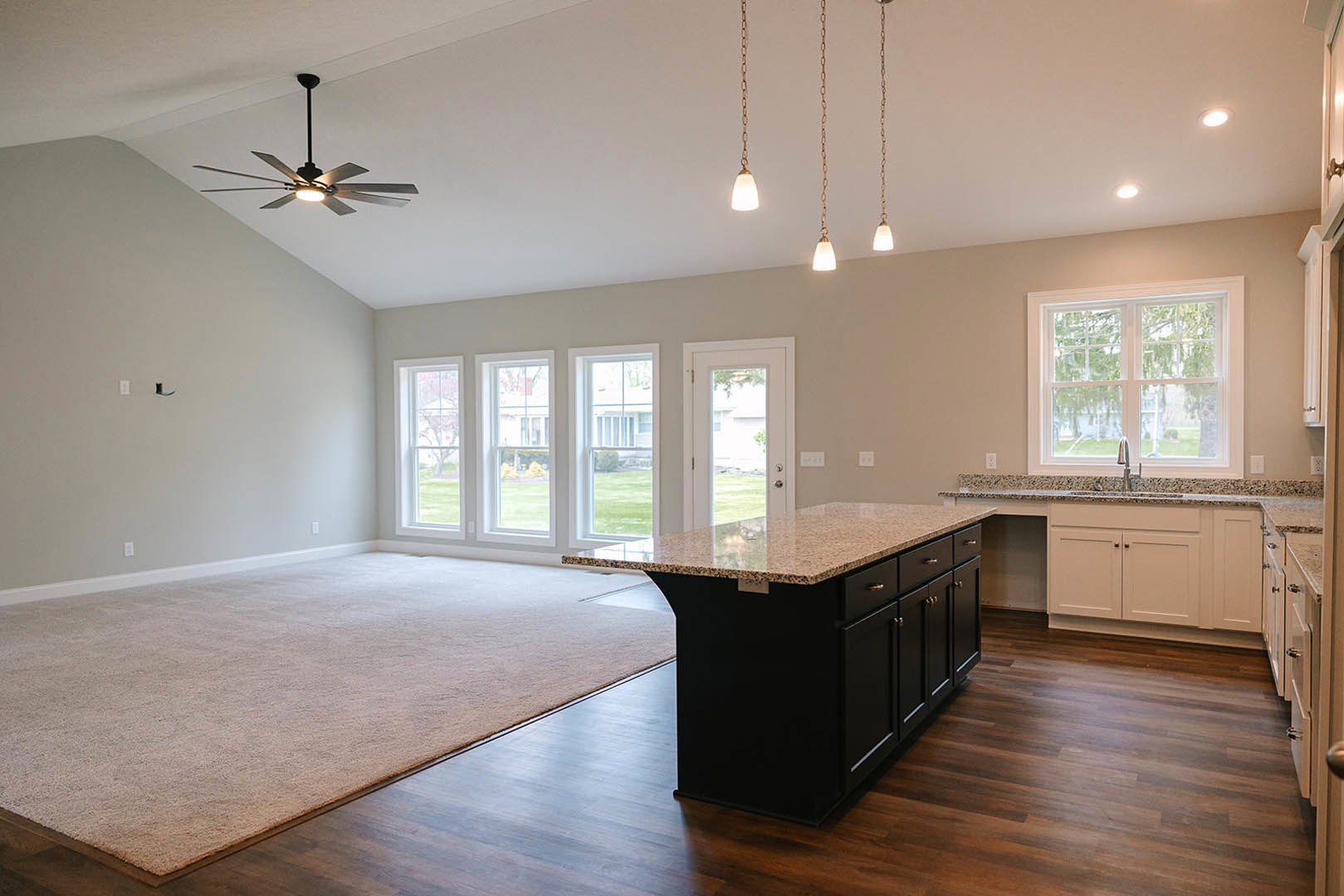 Open-concept kitchen and living room featuring a ceiling fan with light, granite kitchen island, wood cabinetry, carpeted living area, and large windows overlooking trees and a