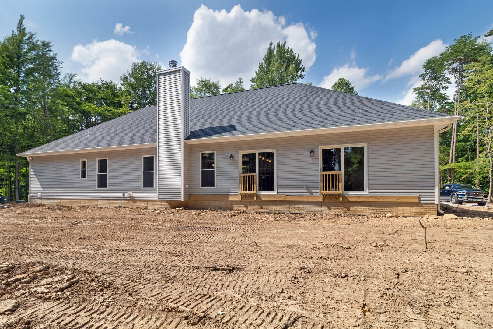 Two-story house with covered porch and brick chimney, dirt yard with tire tracks, barred wooden window, white rectangular vent, overcast sky, and surrounding trees.