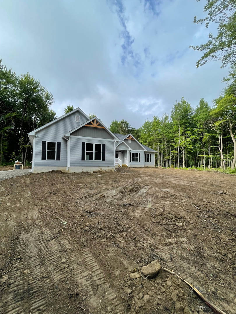 Modern cottage-style house with white-framed windows, prominent roof overhang, surrounded by trees and sky, situated next to a dirt lot with scattered sticks and plants.