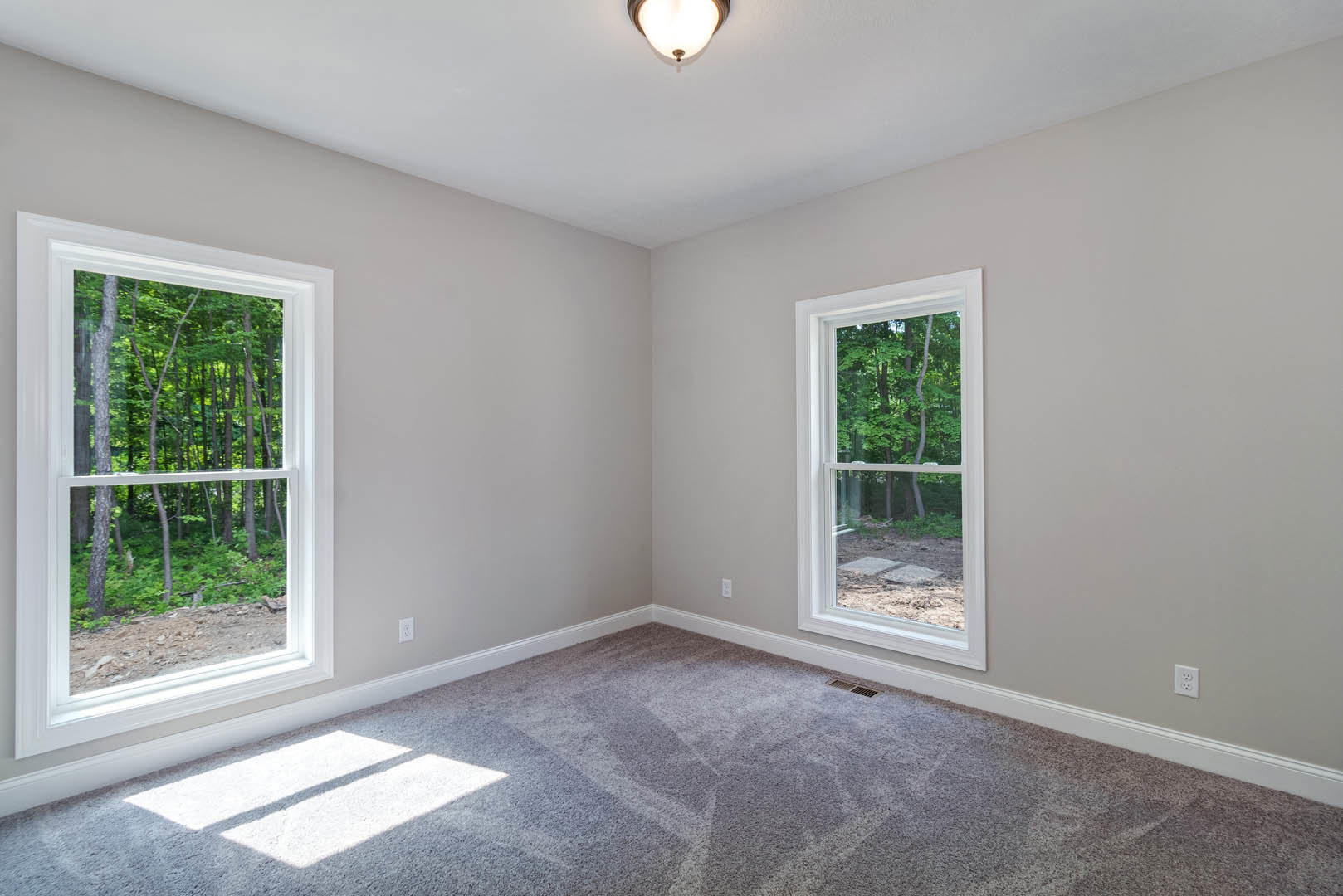 Bright carpeted room with large windows, white walls, ceiling light fixture, and view of green trees outside