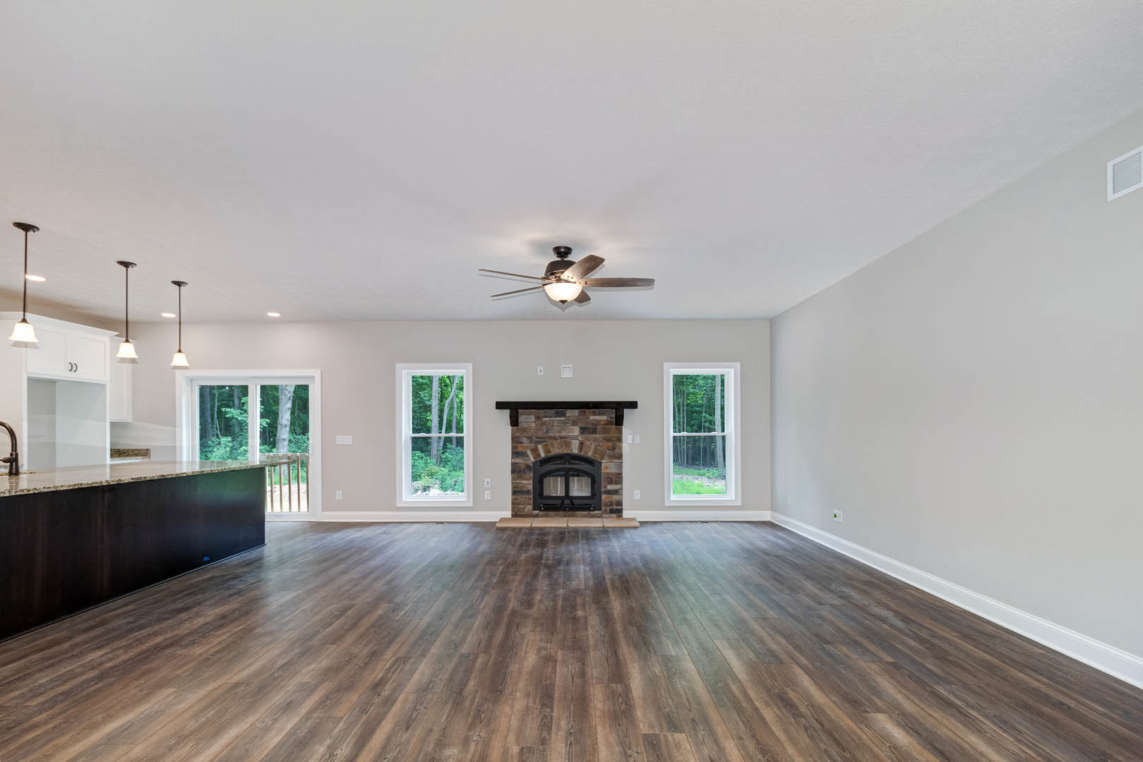 Living room with hardwood floors, white walls, ceiling fan with light, large window overlooking trees, and black-framed fireplace.