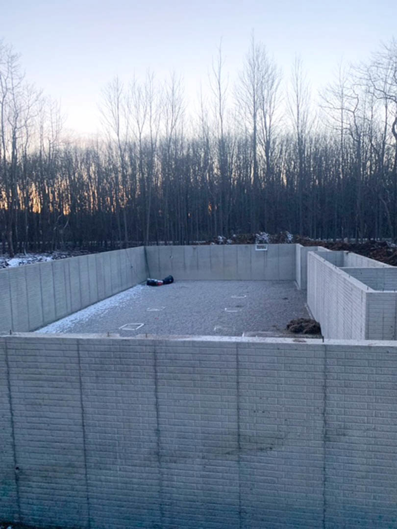 Concrete block wall with rectangular openings, central reflecting pool bordered by snow, leafless trees in background, street light near grey brick wall, white wall with square