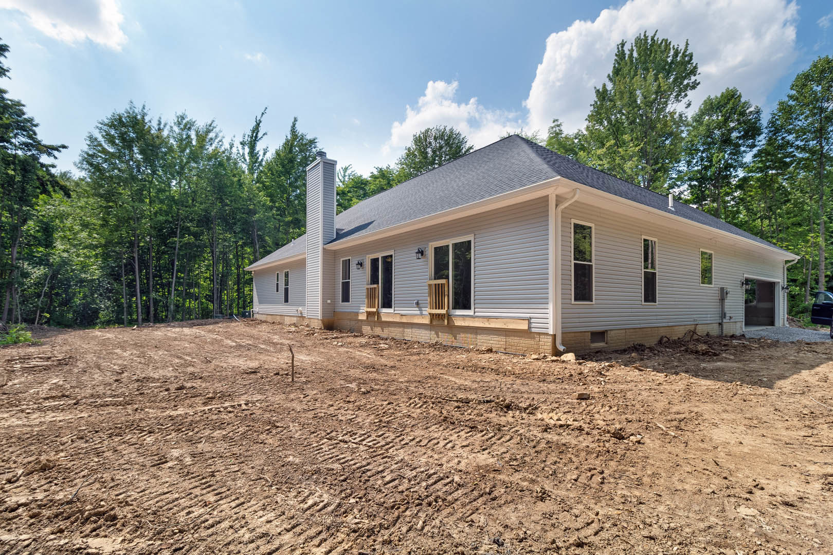 Two-story home under construction with exposed framing, covered porch, large windows, dirt lot with tire tracks, surrounded by mature trees under cloudy sky