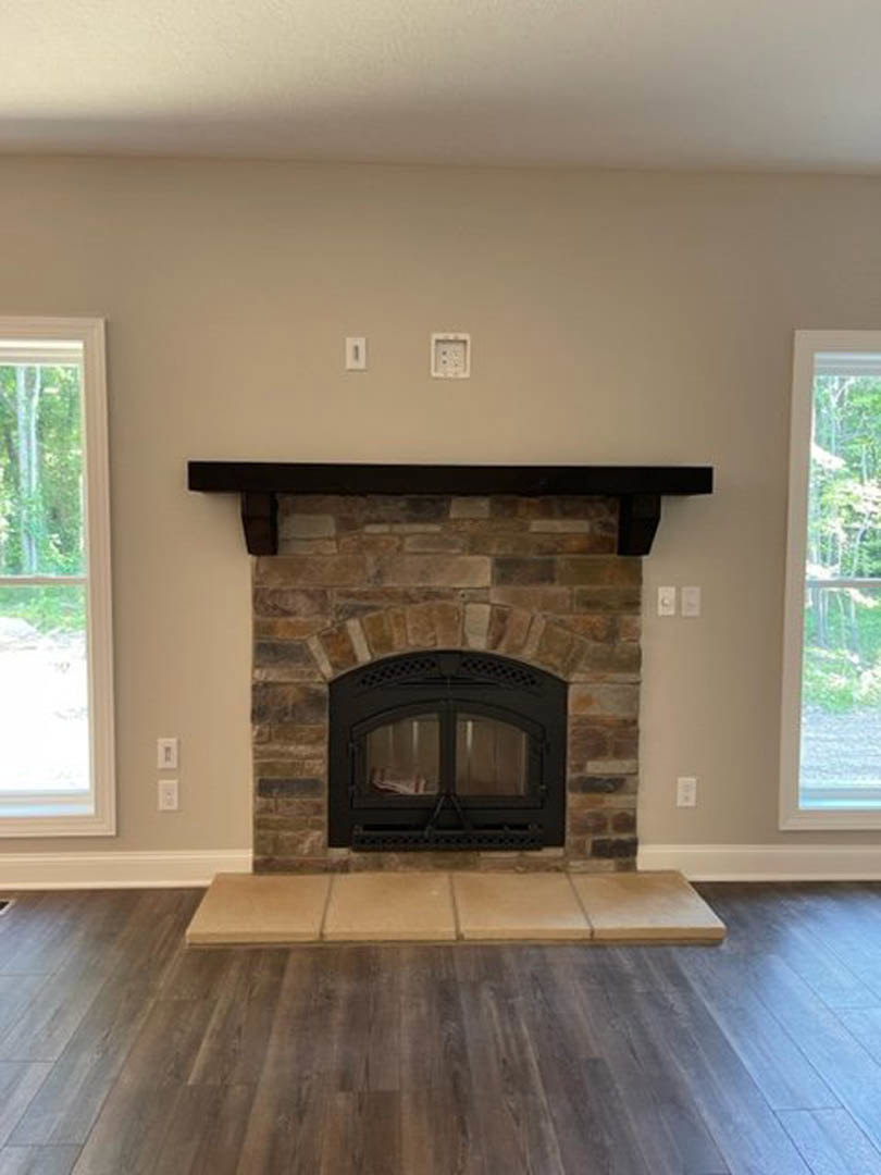 Black-framed glass fireplace set in a tiled wall, wood flooring in foreground, large window with tree views in background