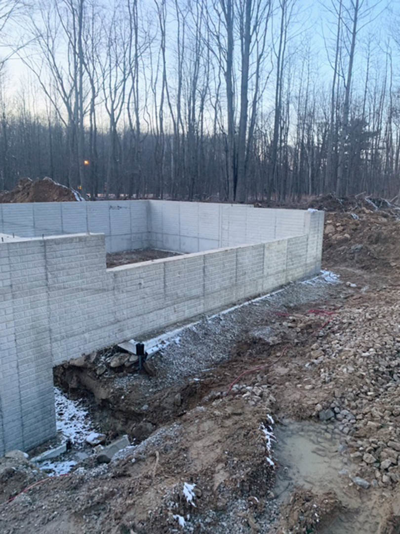 Concrete foundation set in a forest clearing, surrounded by tall trees and exposed dirt, with a partially constructed wall and a hole in the ground.