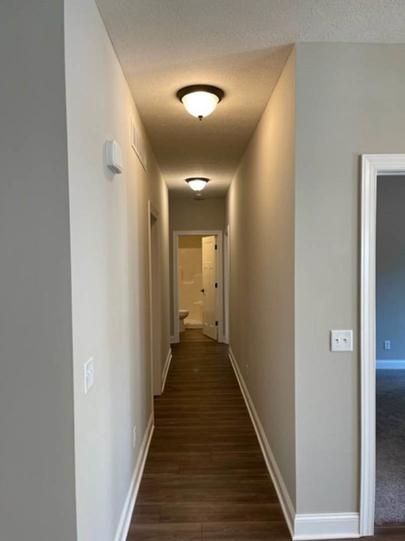 Long hallway with wood flooring, white plaster walls, ceiling light fixture, closed door at the end, and white wall outlet visible on the right.