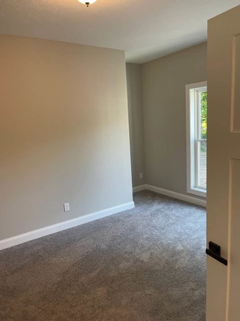 White carpeted floor and white walls in a room with an open door featuring a silver handle; window with white frame visible.
