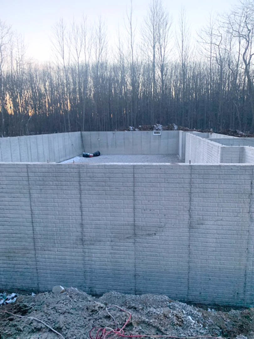 Concrete foundation surrounded by bare trees, with a parked car on the retaining wall, pile of dirt in foreground, red rope visible, and large white wall featuring a central