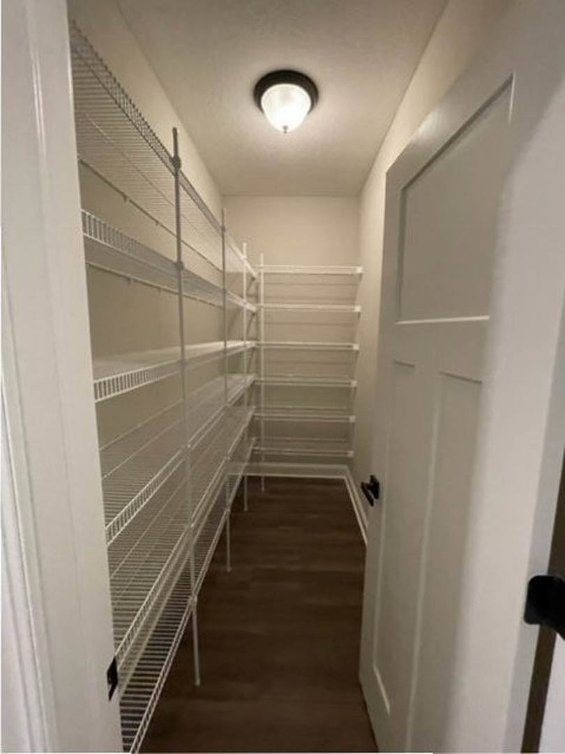 Hallway with white built-in shelves, metal shelf on brown wood floor, ceiling light fixture, plaster walls, door, and staircase visible in background